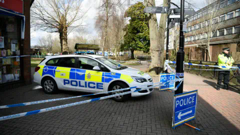 European Photopress Agency A police car behind a police cordon in Salisbury, parked on a brick paved road with parts of buildings visible on either side and treess and a park in the background. A police officers in a yellow high-vis jacket an dflat cap stands to the left hand side of the photo behind the police cordon tape